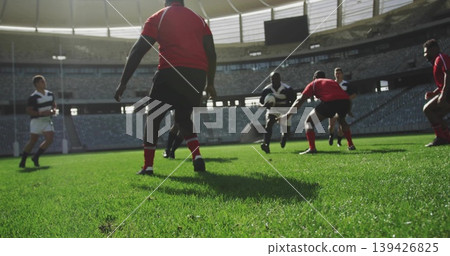 Running striped-jersey player holding rugby ball on stadium grass pitch, with red jerseys tackling Running striped-jersey player holding rugby ball on stadium grass pitch, with red jerseys tackling 139426825