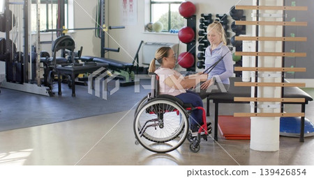 Guiding therapist and wheelchair client holding resistance band in rehab gym, trainer in gray pants Guiding therapist and wheelchair client holding resistance band in rehab gym, trainer in gray pants 139426854