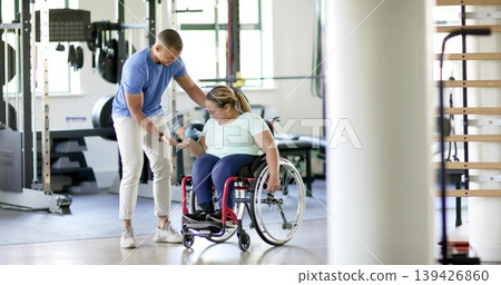 Guiding trainer steadying woman wearing active gear lifting blue dumbbell at gym, red wheelchair Guiding trainer steadying woman wearing active gear lifting blue dumbbell at gym, red wheelchair 139426860