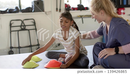 Reaching child sitting crosslegged on padded mat in clinic picking cones with therapist in scrubs Reaching child sitting crosslegged on padded mat in clinic picking cones with therapist in scrubs 139426865