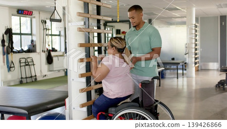 Guiding therapist assisting seated patient in wheelchair in pink tee holding stall bars in clinic Guiding therapist assisting seated patient in wheelchair in pink tee holding stall bars in clinic 139426866