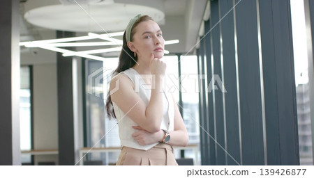 Standing woman resting chin on hand in office near tall windows, wearing white sleeveless top Standing woman resting chin on hand in office near tall windows, wearing white sleeveless top 139426877