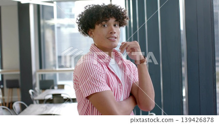 Standing man wearing pink-striped shirt touching chin with wristwatch at coworking lounge, windows Standing man wearing pink-striped shirt touching chin with wristwatch at coworking lounge, windows 139426878