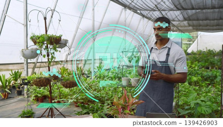 Holding small plant, worker in grey polo and dark apron checking handheld in greenhouse, copy space 139426903