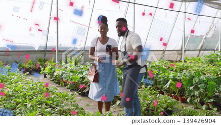 Pointing nursery worker wearing apron guiding buyer in nursery holding phone and bag among plants 139426904