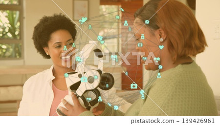 Holding white-black robot, two women in green sweater examining teal AR overlay in living room Holding white-black robot, two women in green sweater examining teal AR overlay in living room 139426961