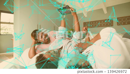 Lying three Black women in loungewear on bed in bright bedroom, holding smartphone, teal overlay 139426972
