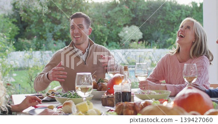 Gesturing diners in tan pullover and pink sweater sharing meal at garden table with wine bottle Gesturing diners in tan pullover and pink sweater sharing meal at garden table with wine bottle 139427060