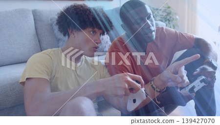 Playing two friends in yellow and rust tees with wireless controllers at home, sunlight streaks Playing two friends in yellow and rust tees with wireless controllers at home, sunlight streaks 139427070