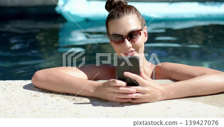 Holding smartphone woman leaning on home pool deck checking phone with dark shades and swim straps 139427076