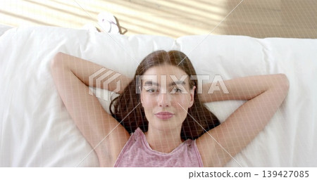Lying woman wearing sleeveless pink top, resting in bedroom on bed with pillow and blind stripes Lying woman wearing sleeveless pink top, resting in bedroom on bed with pillow and blind stripes 139427085