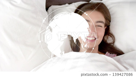 Smiling woman lying in bed under white duvet in bedroom, showing pink neckline and globe overlay Smiling woman lying in bed under white duvet in bedroom, showing pink neckline and globe overlay 139427086