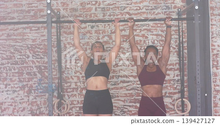 Hanging two women wearing black and burgundy sportswear gripping pull-up bar at gym, rings visible Hanging two women wearing black and burgundy sportswear gripping pull-up bar at gym, rings visible 139427127