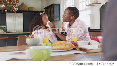 Wiping mother in pink shirt wiping child in heart sweater at wooden kitchen table, with napkin Wiping mother in pink shirt wiping child in heart sweater at wooden kitchen table, with napkin 139427206