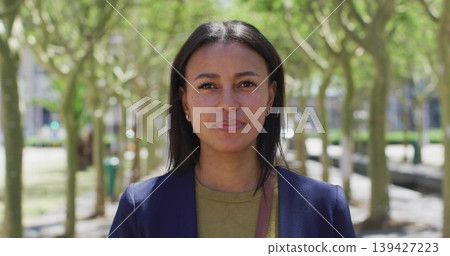 Standing woman wearing navy blazer and olive top in urban park, showing shoulder strap and earrings Standing woman wearing navy blazer and olive top in urban park, showing shoulder strap and earrings 139427223