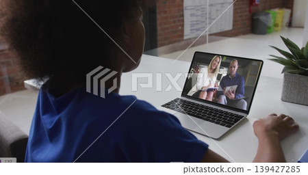 Sitting woman wearing blue top viewing two presenters on laptop at open-plan office desk, succulent 139427285