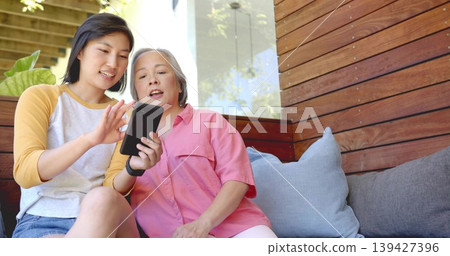 Holding phone, daughter in yellow raglan jean shorts scrolling on porch, mom in pink shirt leaning Holding phone, daughter in yellow raglan jean shorts scrolling on porch, mom in pink shirt leaning 139427396