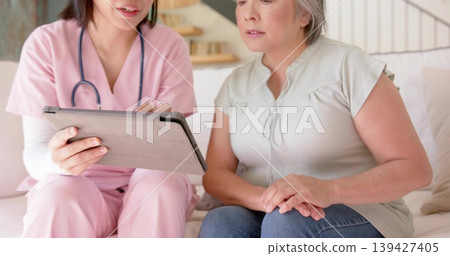 Consulting caregiver in pink scrubs showing tablet to patient in living room, stethoscope visible Consulting caregiver in pink scrubs showing tablet to patient in living room, stethoscope visible 139427405