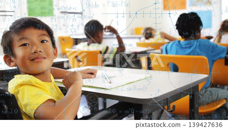Sitting boy wearing yellow T-shirt writing in workbook at elementary classroom, with math overlay Sitting boy wearing yellow T-shirt writing in workbook at elementary classroom, with math overlay 139427636