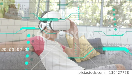 Sitting Asian man wearing VR headset and red-sleeved T-shirt on sofa, interacting with teal HUD Sitting Asian man wearing VR headset and red-sleeved T-shirt on sofa, interacting with teal HUD 139427758
