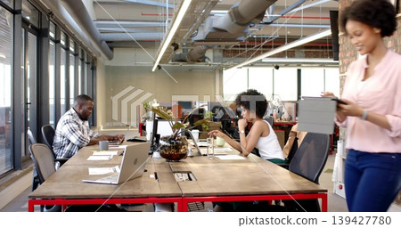 Working three coworkers sharing long table in open office, in casual attire, using laptops, tablet 139427780