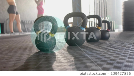Sitting matte black kettlebell with yellow band on gym floor, backlit, women in sportswear standing 139427856