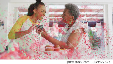 Smiling woman in yellow top holding phone, senior reaching in kitchen, muffins and confetti overlay Smiling woman in yellow top holding phone, senior reaching in kitchen, muffins and confetti overlay 139427873