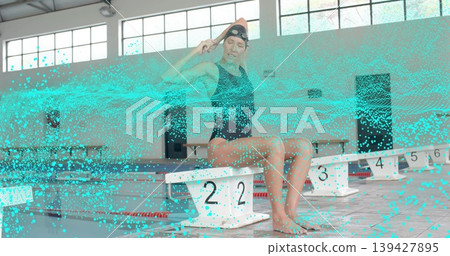 Adjusting cap and goggles, swimmer in black swimsuit sitting on block 2 at pool, turquoise overlay Adjusting cap and goggles, swimmer in black swimsuit sitting on block 2 at pool, turquoise overlay 139427895