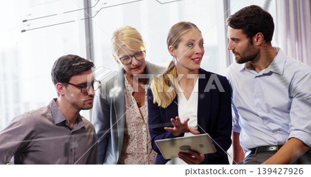 Gesturing presenter and coworkers leaning around tablet by windows, in blazers, reviewing charts Gesturing presenter and coworkers leaning around tablet by windows, in blazers, reviewing charts 139427926