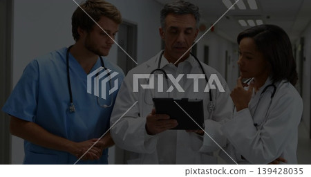 Reviewing medical team studying tablet in hospital corridor, wearing scrubs, coats and stethoscopes Reviewing medical team studying tablet in hospital corridor, wearing scrubs, coats and stethoscopes 139428035