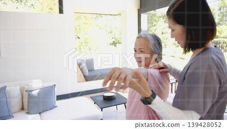 Extending senior in pink-top getting shoulder help from carer-in-scrubs by sofa at home, copy space Extending senior in pink-top getting shoulder help from carer-in-scrubs by sofa at home, copy space 139428052