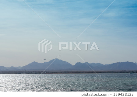 Tranquil seascape with distant mountains and airplane under clear sky Tranquil seascape with distant mountains and airplane under clear sky 139428122