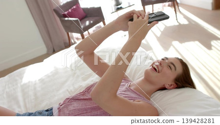 Laughing woman wearing pink top and denim shorts holding phone overhead on white duvet in bedroom Laughing woman wearing pink top and denim shorts holding phone overhead on white duvet in bedroom 139428281