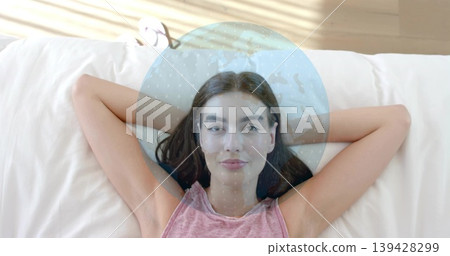 Reclining woman wearing pink sleeveless top on bed at home, with round pillow and globe overlay 139428299