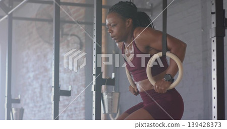 Holding woman wearing maroon sports bra performing ring dips on wooden rings in gym, copy space Holding woman wearing maroon sports bra performing ring dips on wooden rings in gym, copy space 139428373