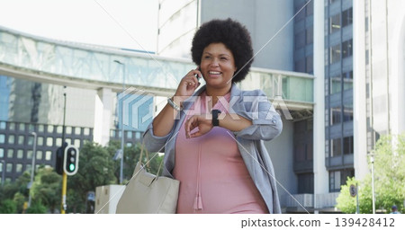 Smiling professional woman checking smartwatch and talking on phone by office towers, blazer Smiling professional woman checking smartwatch and talking on phone by office towers, blazer 139428412