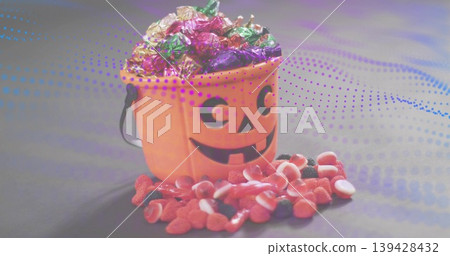 Displaying orange jack-o-lantern bucket holding foil-wrapped sweets on gray table, spilling gummies Displaying orange jack-o-lantern bucket holding foil-wrapped sweets on gray table, spilling gummies 139428432