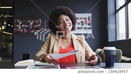 Smiling African American woman in beige blazer handing printed pages at office table with notebook Smiling African American woman in beige blazer handing printed pages at office table with notebook 139428457
