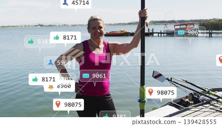 Standing mature rower wearing pink top, black shorts on dock, holding oar by shell, social icons Standing mature rower wearing pink top, black shorts on dock, holding oar by shell, social icons 139428555