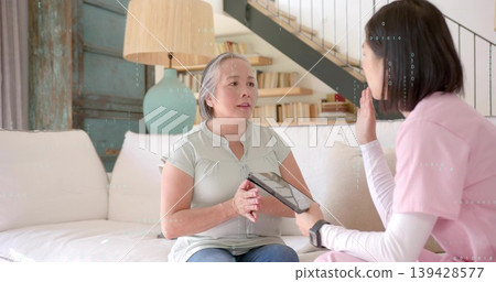 Senior woman and caregiver in pink scrubs discussing care on sofa, caregiver holding tablet 139428577
