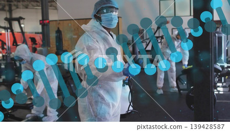 Spraying female worker wearing full PPE disinfecting fitness center with fogger, DNA helix overlay Spraying female worker wearing full PPE disinfecting fitness center with fogger, DNA helix overlay 139428587
