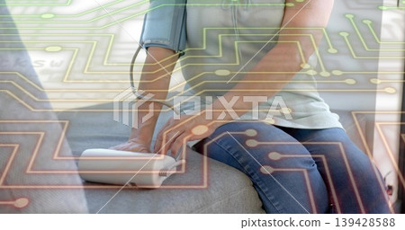 Sitting woman green-top measuring arm-cuff at home on grey-sofa with white-monitor and circuitry Sitting woman green-top measuring arm-cuff at home on grey-sofa with white-monitor and circuitry 139428588