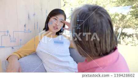 Talking Korean woman in shirt touching temple, leaning on gray couch on porch, blue circuit overlay Talking Korean woman in shirt touching temple, leaning on gray couch on porch, blue circuit overlay 139428602