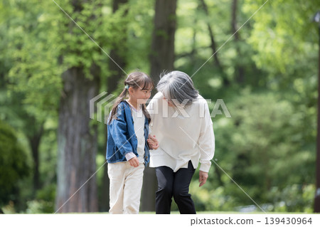 A grandmother plays in a park surrounded by fresh spring greenery, while her smiling granddaughter walks beside her, taking care of her grandchild. 139430964