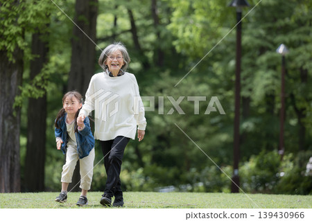 A full-body shot of a grandmother playing in a park amidst the fresh greenery of spring, her granddaughter walking hand-in-hand with her, her face beaming and looking directly at the camera. (Copy space on the right) 139430966