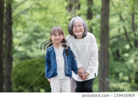 A close-up shot of a grandmother holding hands and her smiling granddaughter playing in a park amidst the fresh greenery of spring, looking directly at the camera. 139430969