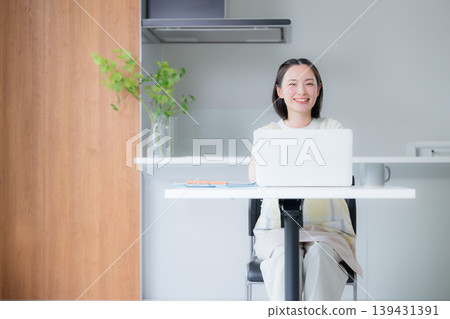 A smiling woman working on her laptop in the dining room; a remote worker; a woman working from home or studying for qualifications at home. A smiling woman working on her laptop in the dining room; a remote worker; a woman working from home or studying for qualifications at home. 139431391