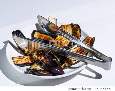 Grilled eggplant slices in a white bowl with metal tongs 139431660