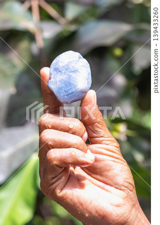 Gemstones in Sri Lanka. Close-up of a human hand holding a smooth, light blue irregular stone, sparkling with water droplets against a soft green foliage background. Sri Lanka island in Asia. 139433260