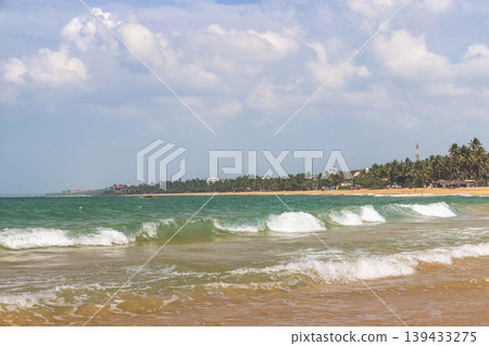 Vibrant Tropical Beach Scene with Rolling Turquoise Waves and Distant Palm Tree Lined Coastline Under a Cloudy Sky, Serene Tropical Shoreline Landscape. Sri Lanka island in Asia. Vibrant Tropical Beach Scene with Rolling Turquoise Waves and Distant Palm Tree Lined Coastline Under a Cloudy Sky, Serene Tropical Shoreline Landscape. Sri Lanka island in Asia. 139433275